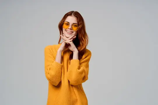 Рыжеволосая девушка в желтых очках и свитере улыбается на сером фоне