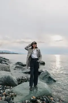 Молодая женщина в плаще и кепке стоит на камнях у моря