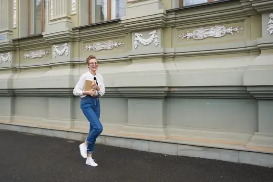 Улыбчивая девушка в очках с блокнотом позирует у фасада здания