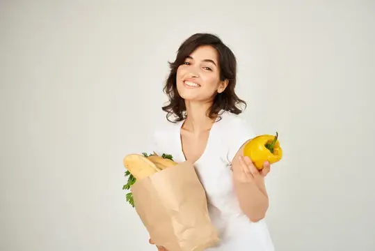 Улыбающаяся брюнетка с бумажным пакетом продуктов и жёлтым перцем