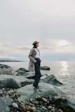 Женщина в плаще и кепке стоит на камнях у моря в пасмурный день