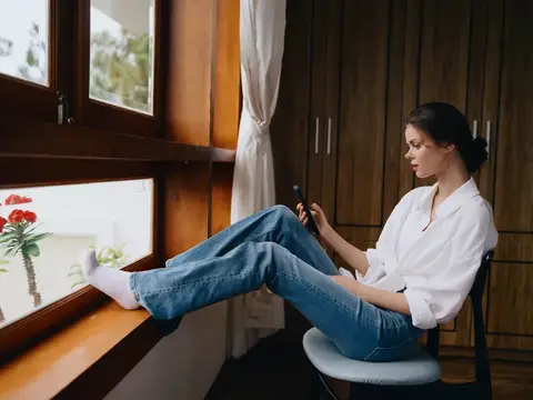 Молодая женщина в белой рубашке сидит у окна с телефоном