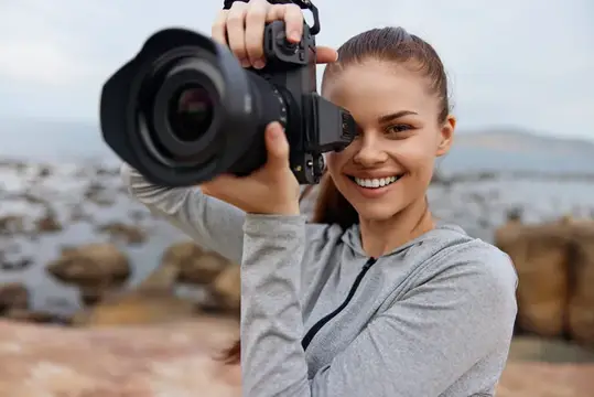 Улыбающаяся девушка смотрит в видоискатель камеры на каменистом берегу