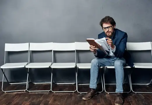 Мужчина в очках читает книгу на белом стуле у серой стены
