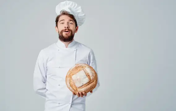 Мужчина в сером фартуке и головном уборе жестикулирует руками как профессиональный повар