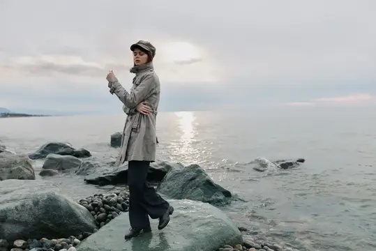 Мужчина в плаще и кепке стоит на камнях у моря в пасмурный день