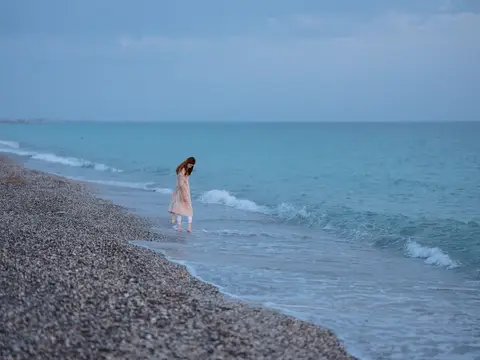 Девушка в светлом платье идёт по кромке воды у галечного берега