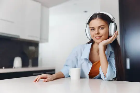 Молодая женщина наслаждается музыкой в наушниках, находясь на кухне дома, чувствуя себя расслабленной и развлечённой