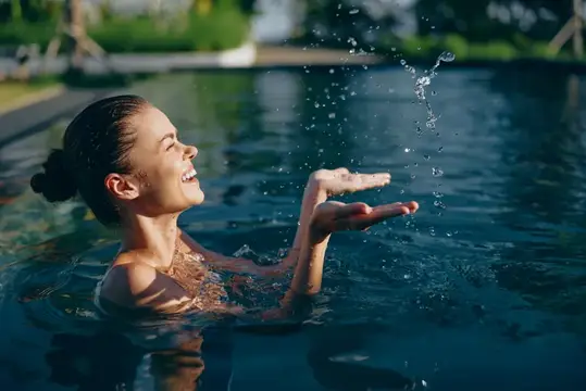 Счастливая женщина в плавательном бассейне играет с брызгами воды, подняв руки вверх, улыбаясь с закрытыми глазами и наслаждаясь летним моментом активного отдыха на свежем воздухе