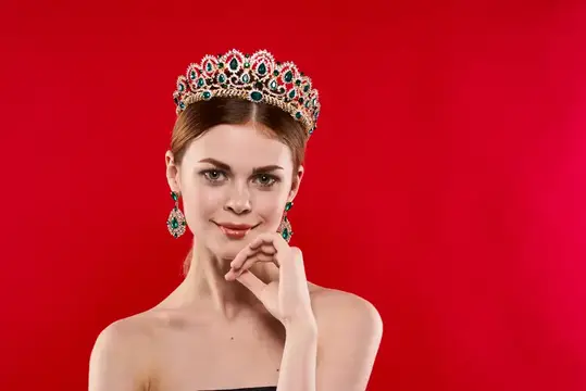Красивая женщина с обнаженными плечами, короной на голове, косметикой на красном фоне