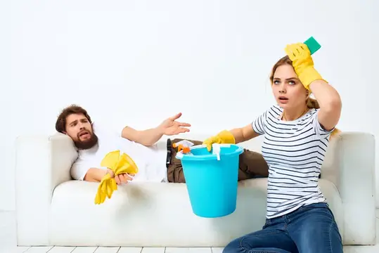 Уставшая женщина убирается, пока мужчина отдыхает на диване