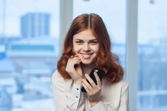 Рыжеволосая девушка улыбается с телефоном в руке у окна