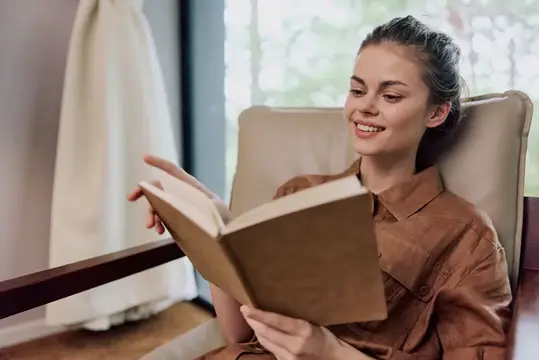 Улыбающаяся девушка с темными волосами читает книгу в кресле у окна