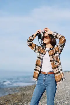 Улыбающаяся девушка в клетчатой рубашке фотографирует на камеру у моря
