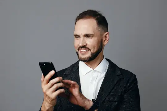 Улыбающийся бородатый мужчина в пиджаке смотрит в смартфон