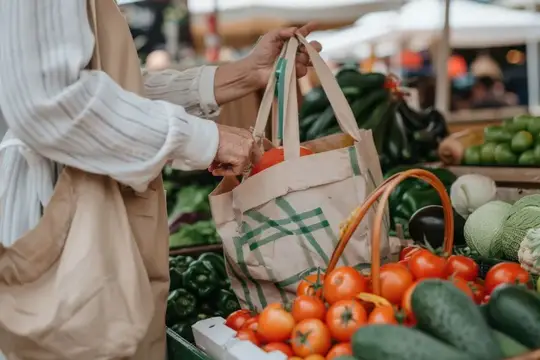 Женщина покупает свежие фрукты и овощи на фермерском рынке, держа продуктовый пакет