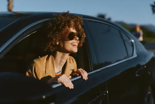 Кудрявая девушка в солнечных очках улыбается, высунувшись из окна автомобиля