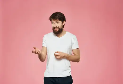 Бородатый мужчина в белой футболке жестикулирует на розовом фоне
