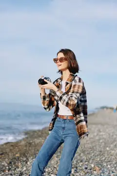 Молодая женщина в клетчатой рубашке фотографирует на галечном пляже
