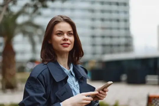 Яркий непринужденный портрет улыбающейся женщины на улице со смартфоном, городская обстановка, повседневная одежда, джинсовая куртка, светлая рубашка, иллюстрирующий современную связь и повседневную…
