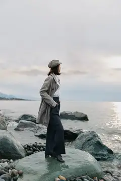 Молодая женщина в плаще и кепке стоит на камнях у моря