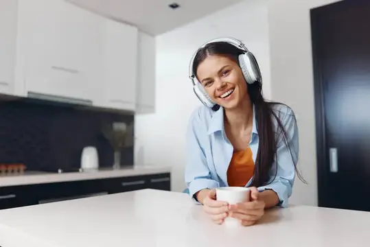 Счастливая молодая женщина наслаждается музыкой в наушниках и кофе у кухонной стойки