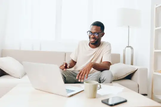 Улыбающийся темнокожий мужчина в очках работает за ноутбуком на диване