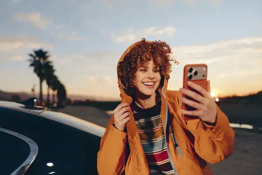 Кудрявая девушка в оранжевой куртке делает селфи на закате