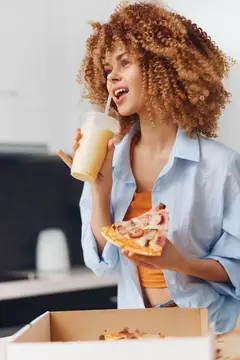 Кудрявая девушка пьёт смузи и ест пиццу на кухне