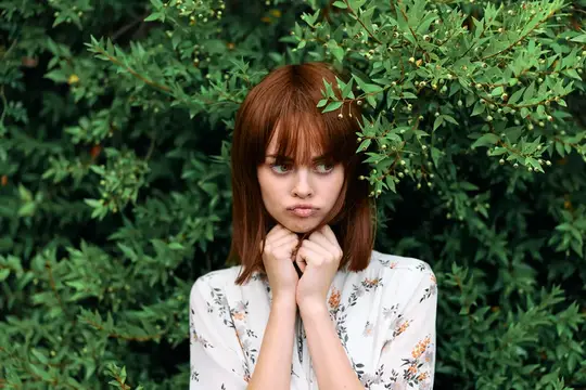 Рыжеволосая девушка с каре стоит среди зелёных кустов