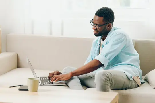 Улыбающийся афроамериканский мужчина работает за ноутбуком в современной гостиной, совершает звонок для онлайн-покупок, в окружении удобного дивана и очков