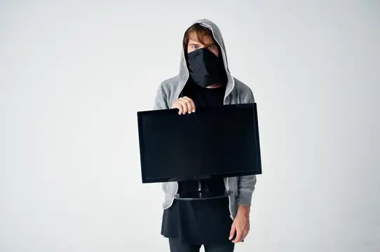 Мужчина в капюшоне и маске держит монитор на белом фоне