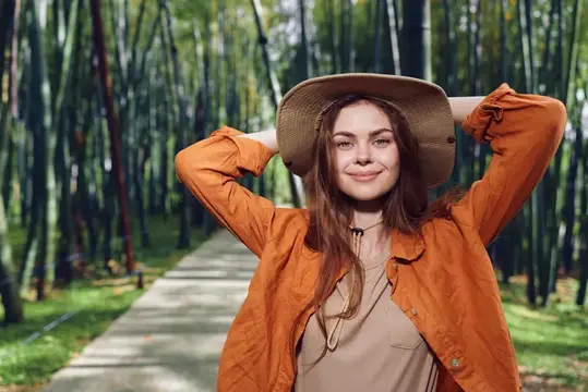 Улыбающаяся девушка в оранжевой куртке и шляпе на фоне бамбукового леса