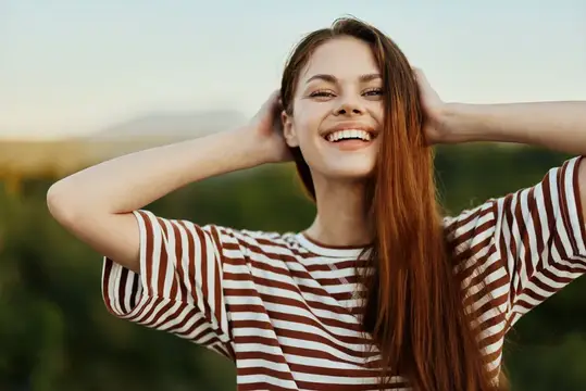 Улыбающаяся девушка с длинными волосами поправляет причёску на природе