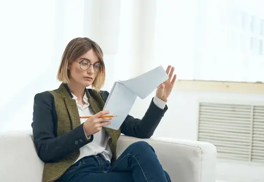 Молодая женщина в очках с блокнотом сидит на диване у окна