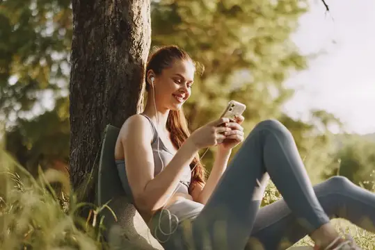 Девушка в спортивной форме сидит под деревом со смартфоном и наушниками