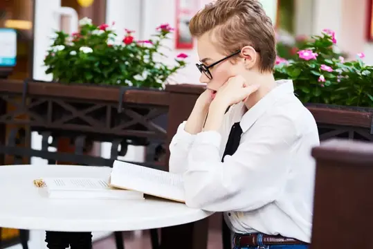 Рыжеволосый мальчик в очках читает книгу за столиком летнего кафе