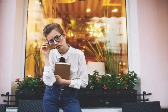 Образованная женщина с книгой и в очках сидит в кресле на веранде кафе