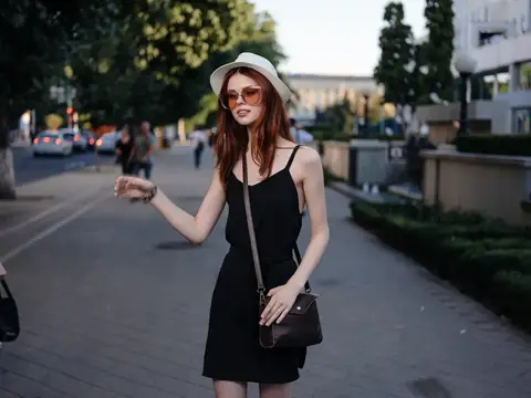 Рыжеволосая девушка в шляпе и солнечных очках идёт по летней улице