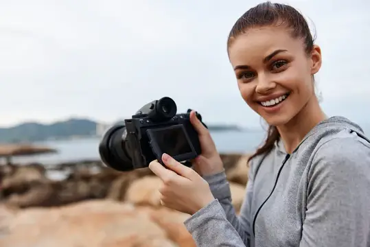 Улыбающаяся женщина-фотограф на каменистом берегу, запечатлевающая моменты с помощью фотоаппарата, излучающая радость и энтузиазм по поводу уличной фотографии, природы и приключений