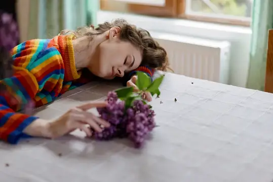Интимная домашняя обстановка: женщина в красочном свитере засыпает рядом с цветами, что подчеркивает отдых, заботу о себе и нежную, повседневную уязвимость в уютном пространстве
