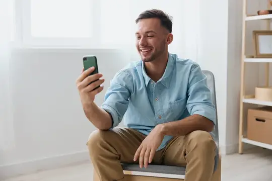 Улыбающийся мужчина в голубой рубашке смотрит в смартфон дома