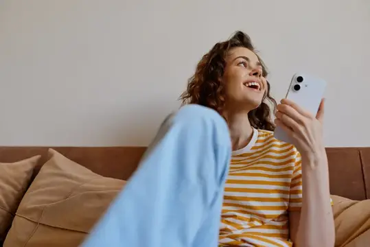 Кудрявая девушка в полосатой футболке смеётся с телефоном на диване