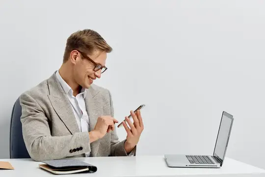 Улыбающийся мужчина в очках смотрит в смартфон за рабочим столом