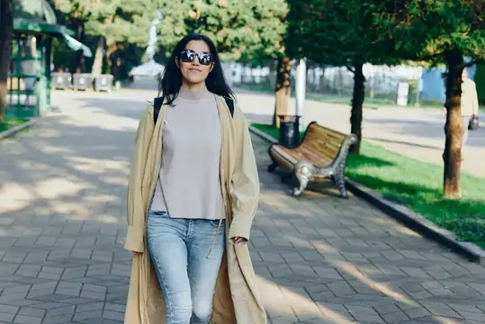 Улыбающаяся темноволосая девушка в солнцезащитных очках гуляет по аллее парка