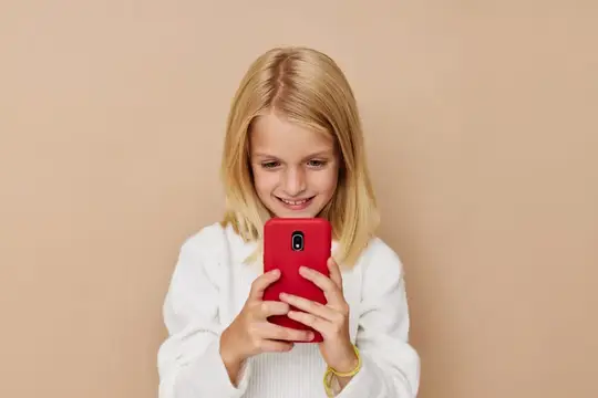 Светловолосая девочка улыбается, глядя в красный смартфон в руках