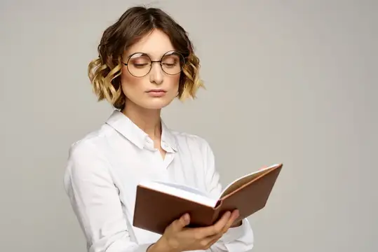 Молодая женщина в очках и белой рубашке читает книгу на сером фоне