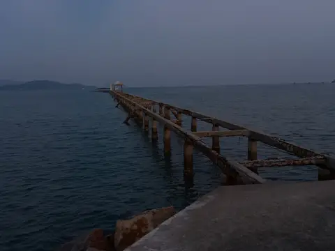 Ржавый старый пирс, уходящий в спокойные морские воды в сумерках, с глубокими синими тонами и видимыми вдалеке островами, создающий мирную и безмятежную прибрежную сцену