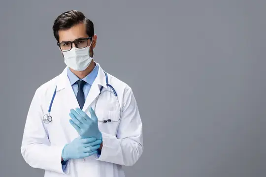 Врач в маске и перчатках поправляет халат на сером фоне