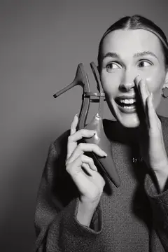 Улыбающаяся светлокожая женщина-европейка держит туфлю возле лица, шепча с волнением, черно-белое фото на студийном фоне